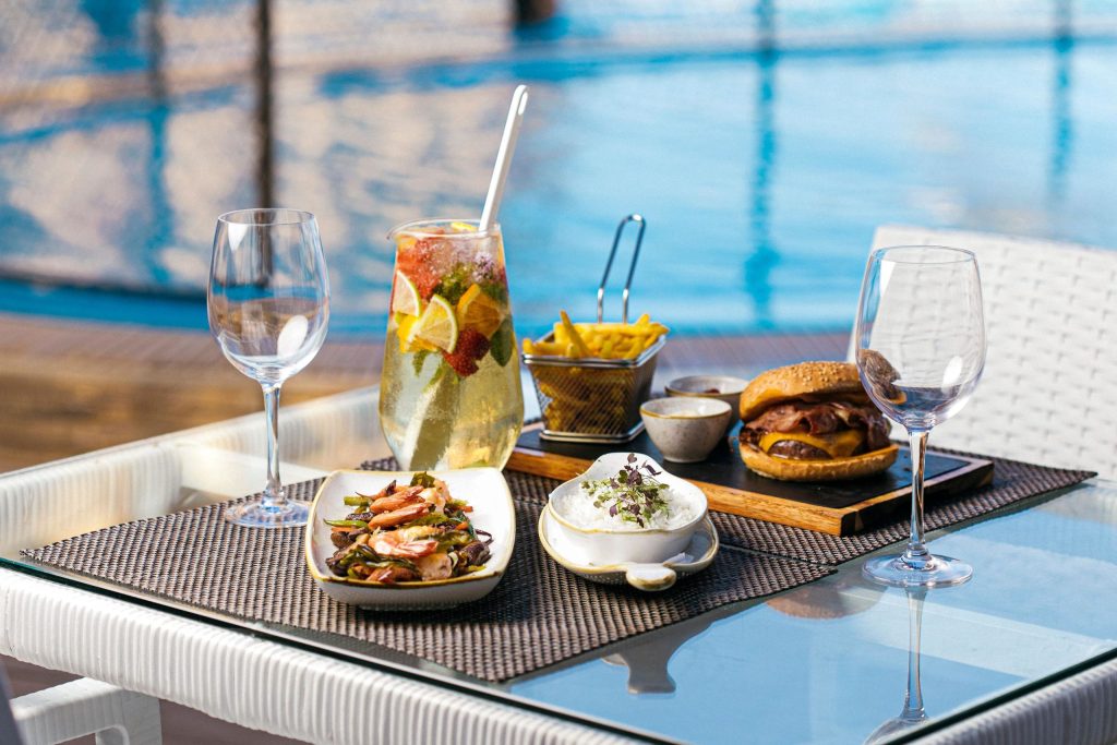 Outdoor table with a burger, fries, rice, seafood, and a fruity drink by a pool.