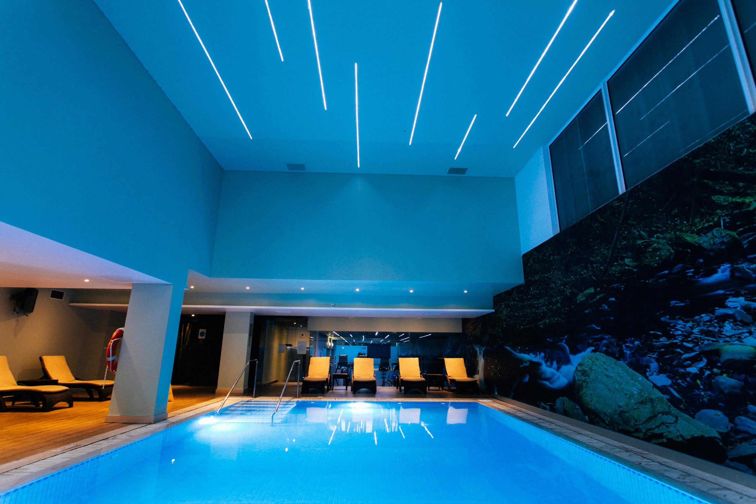 Indoor pool with lounge chairs, modern lighting, and nature-themed wall decor.