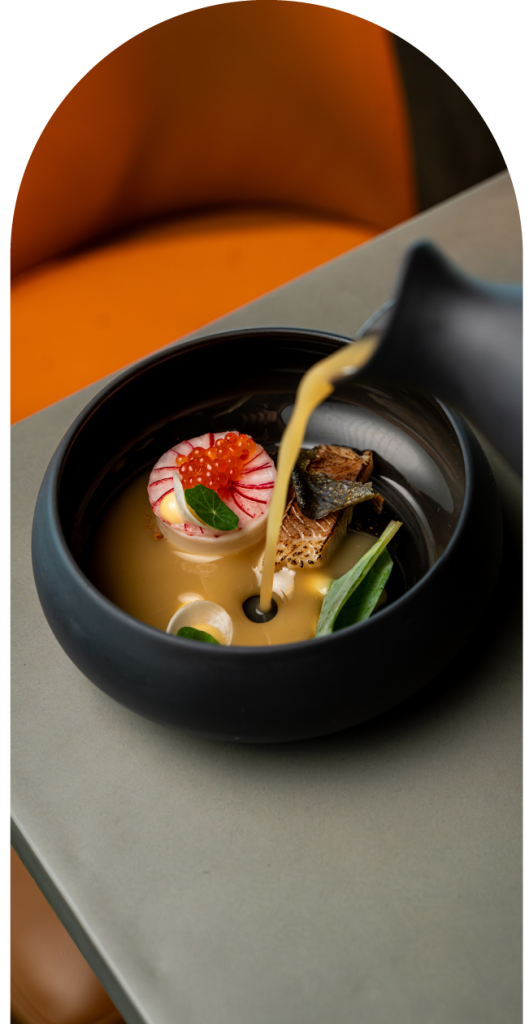 A black bowl with food garnished with roe, greens, and a sauce being poured.