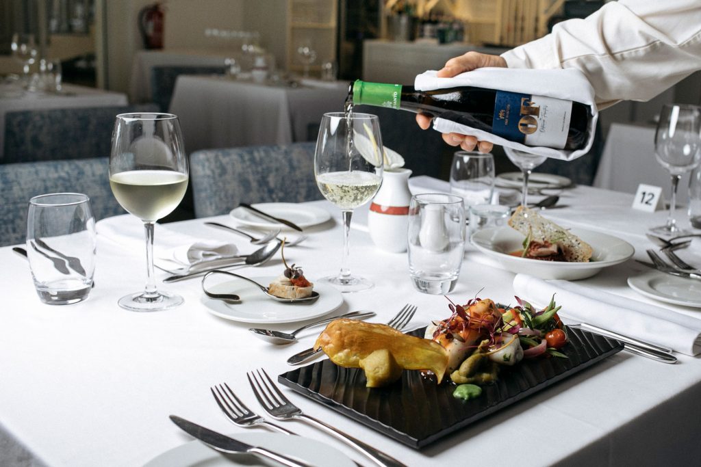 Fine dining table with white wine being poured, gourmet dishes, and elegant setting.