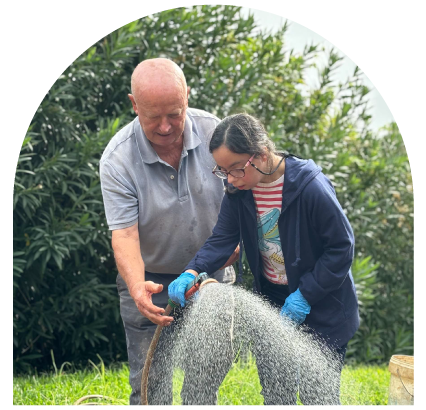 Older man helps girl water plants with a hose in a garden.
