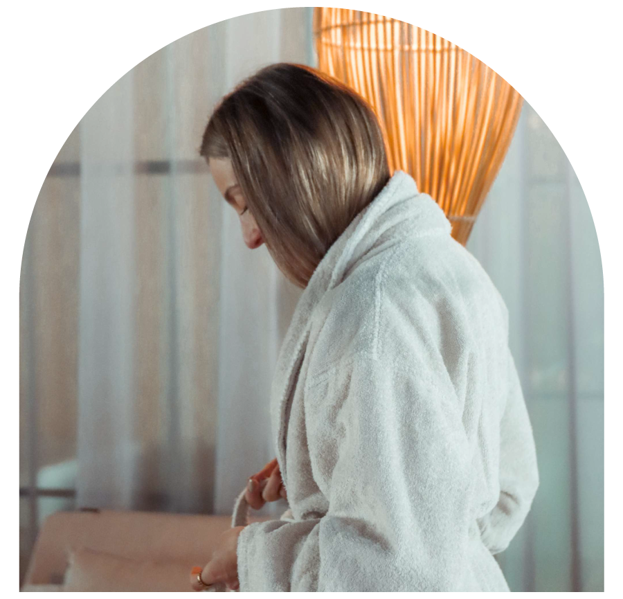 Woman in a bathrobe standing near a warm lamp.