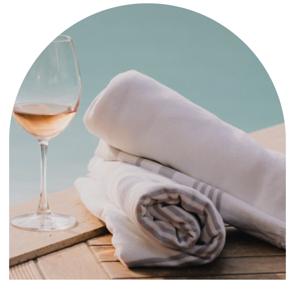 Glass of wine and rolled towels on a wooden table by a pool.