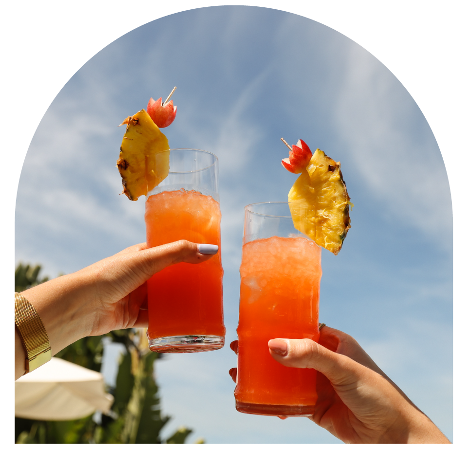 Two hands hold fruit cocktails with pineapple garnishes against a blue sky.