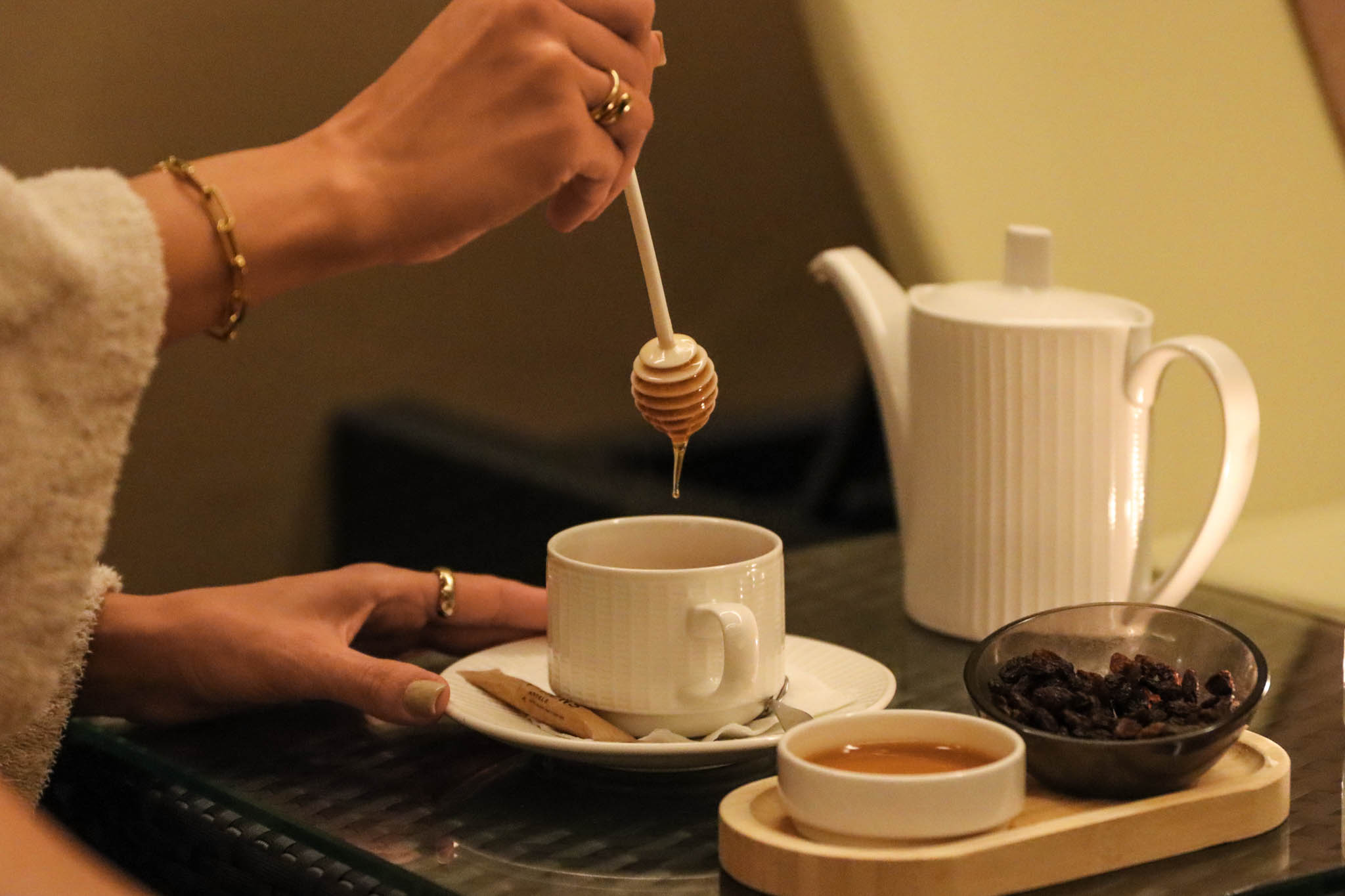 Hand drizzles honey into a cup of tea; teapot and snacks on the side.