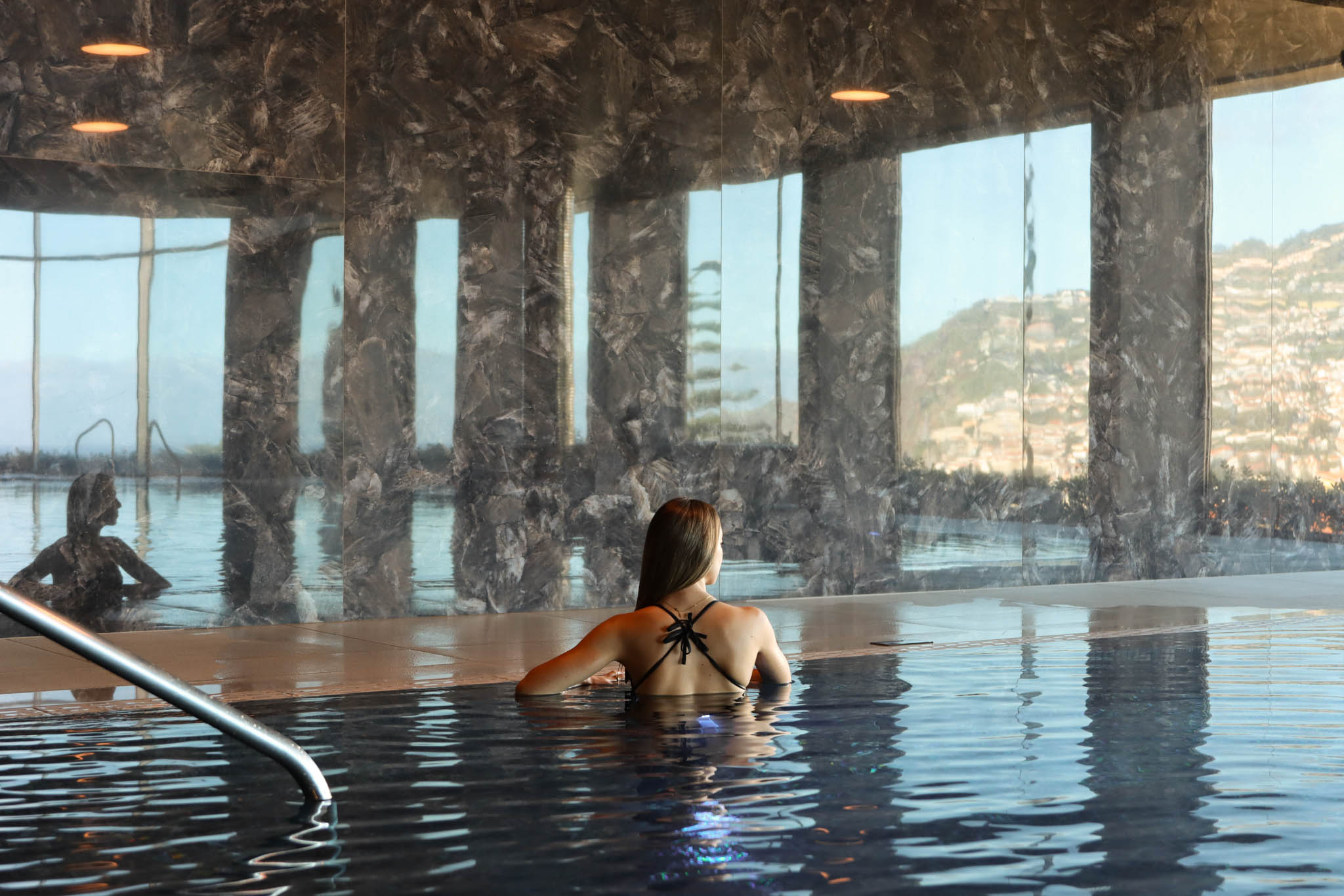Woman in a pool indoors with mountain view through large windows.