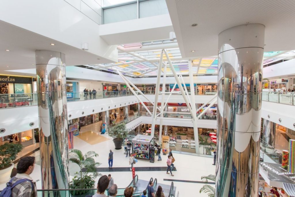 Shopping mall interior with escalators, shoppers, and colorful glass ceiling.