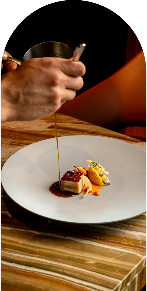 Hand pouring sauce over a gourmet dish with meat and vegetables on a white plate.