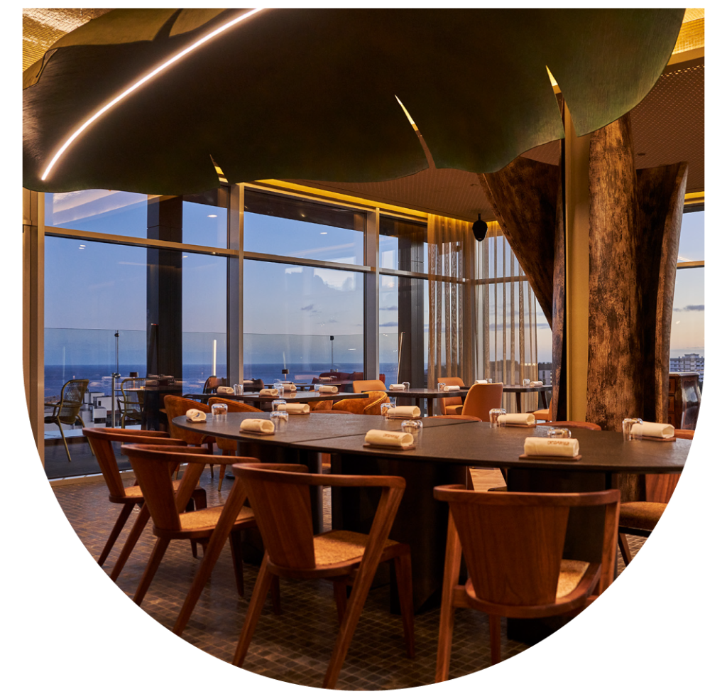 Modern dining area with wooden chairs, tree sculptures, and ocean view through large windows.