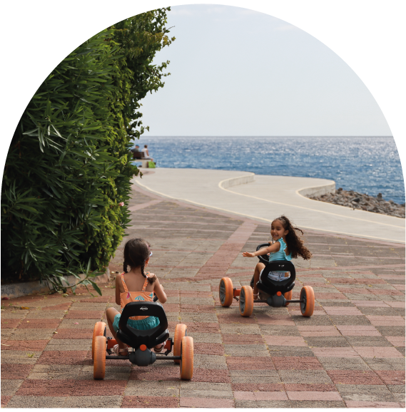 Two children ride toy cars on a seaside path under a clear sky.