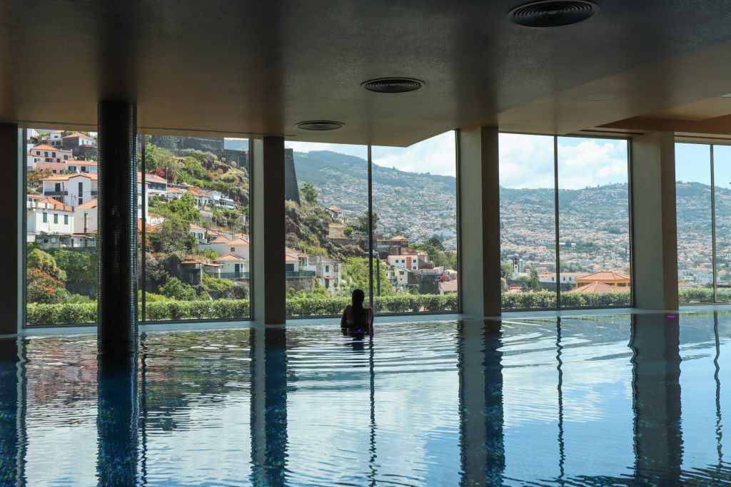 Person sitting in indoor pool with a scenic view of hills and houses through large windows.
