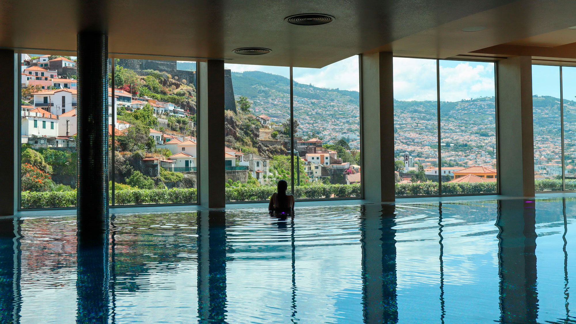 Person in indoor pool with scenic hillside view through large windows.