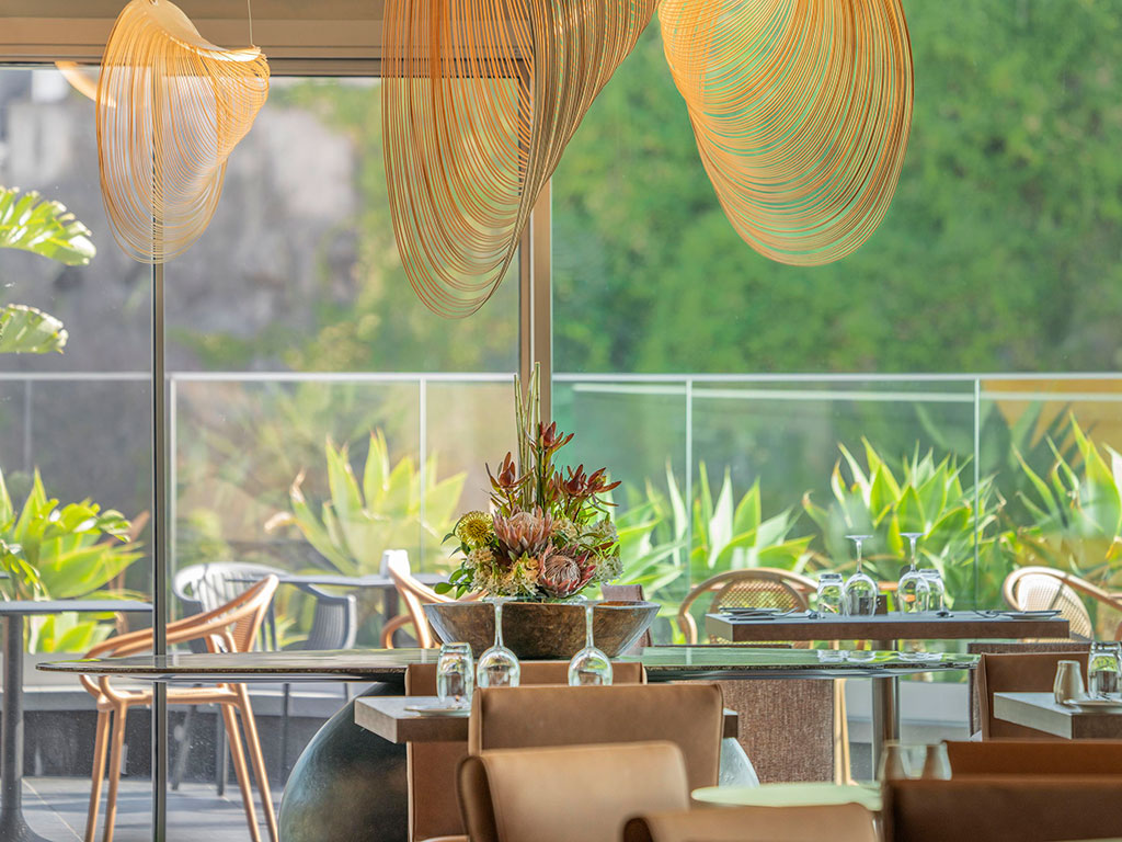 Modern restaurant with stylish lighting and lush greenery outside large windows.