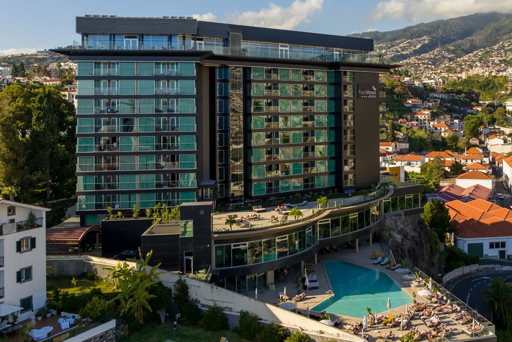 Modern hotel with a pool, set in a cityscape with hills in the background.