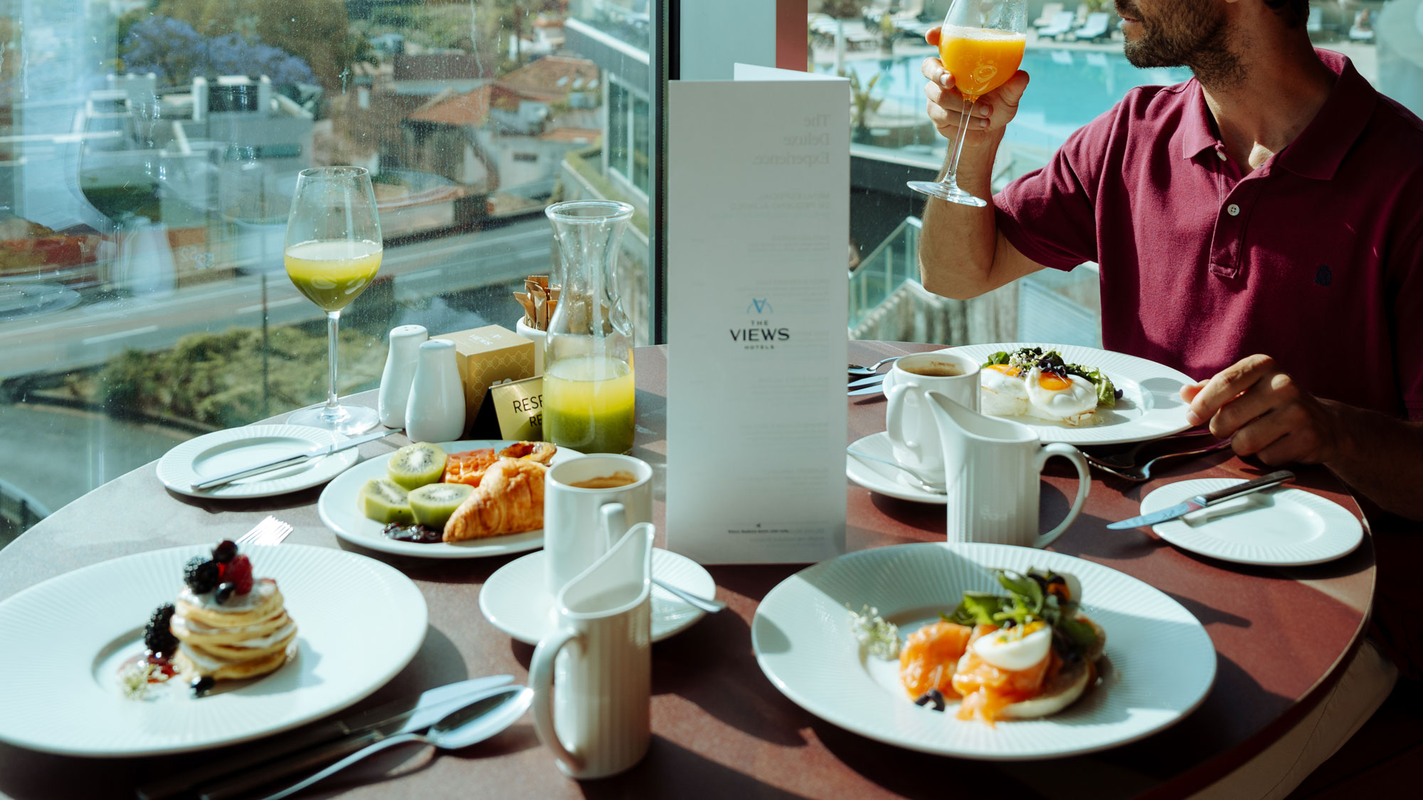 A man having breakfast by a window with juice, pancakes, and a croissant on the table.