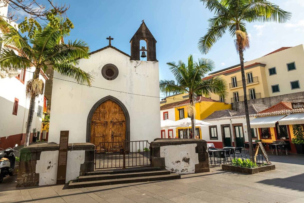 A small white chapel with wooden doors, flanked by palm trees, under a clear blue sky.