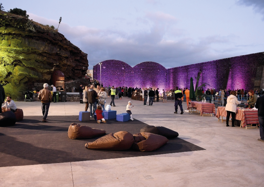 Crowd at an outdoor event with purple-lit walls, beanbags, and tables under a cloudy sky.