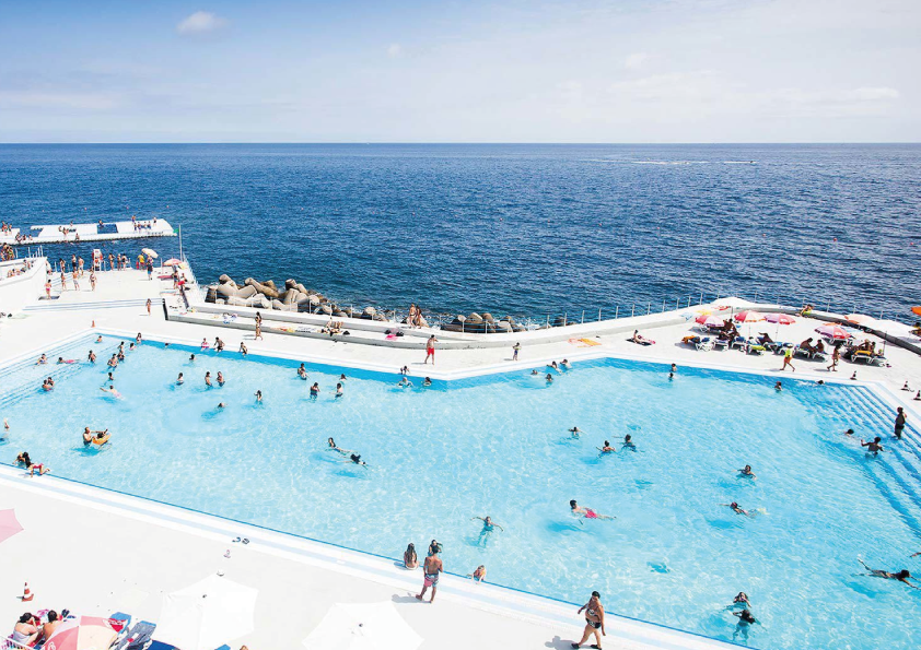 A seaside pool with people swimming, sunbathing, and relaxing under colorful umbrellas.