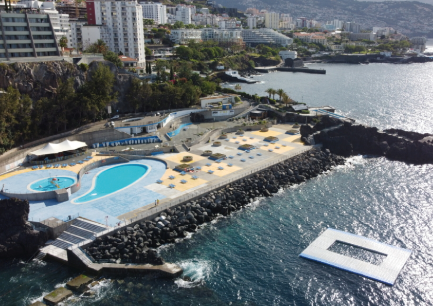 Coastal view of a swimming pool complex beside the ocean with city and hills in the background.