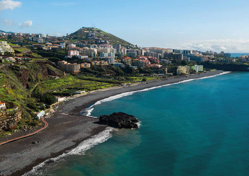 Coastal cityscape with a mountain, buildings, and a rocky beach along a blue ocean.