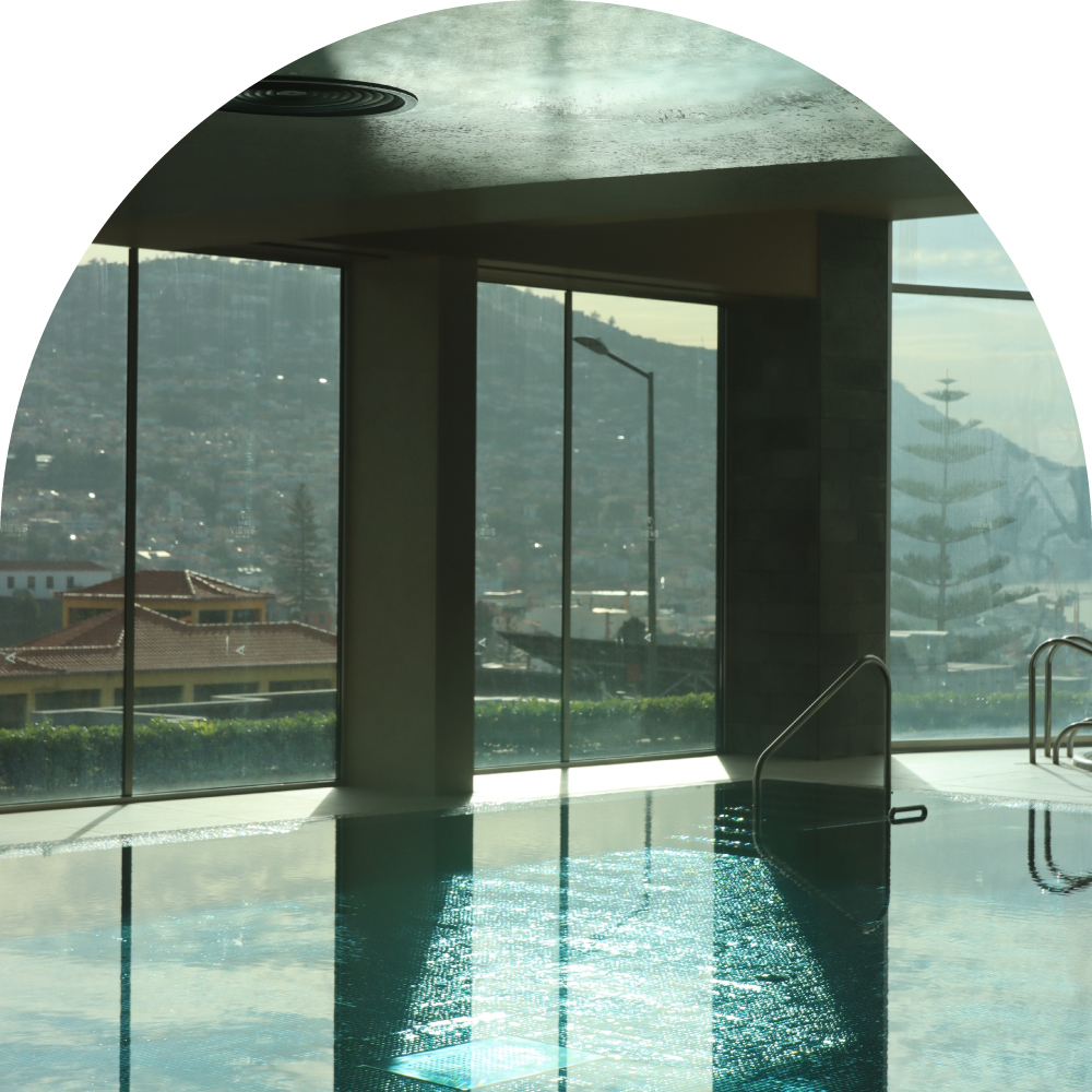 Indoor pool with city and mountain view through large arched glass windows.