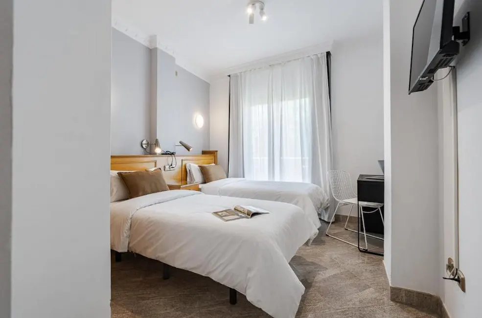 Habitaci&oacute;n con dos camas, ventana luminosa y televisi&oacute;n en la pared.