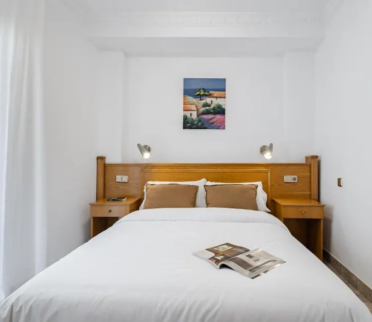 Cama matrimonial con cabecera y cuadros, libro abierto sobre la colcha blanca.