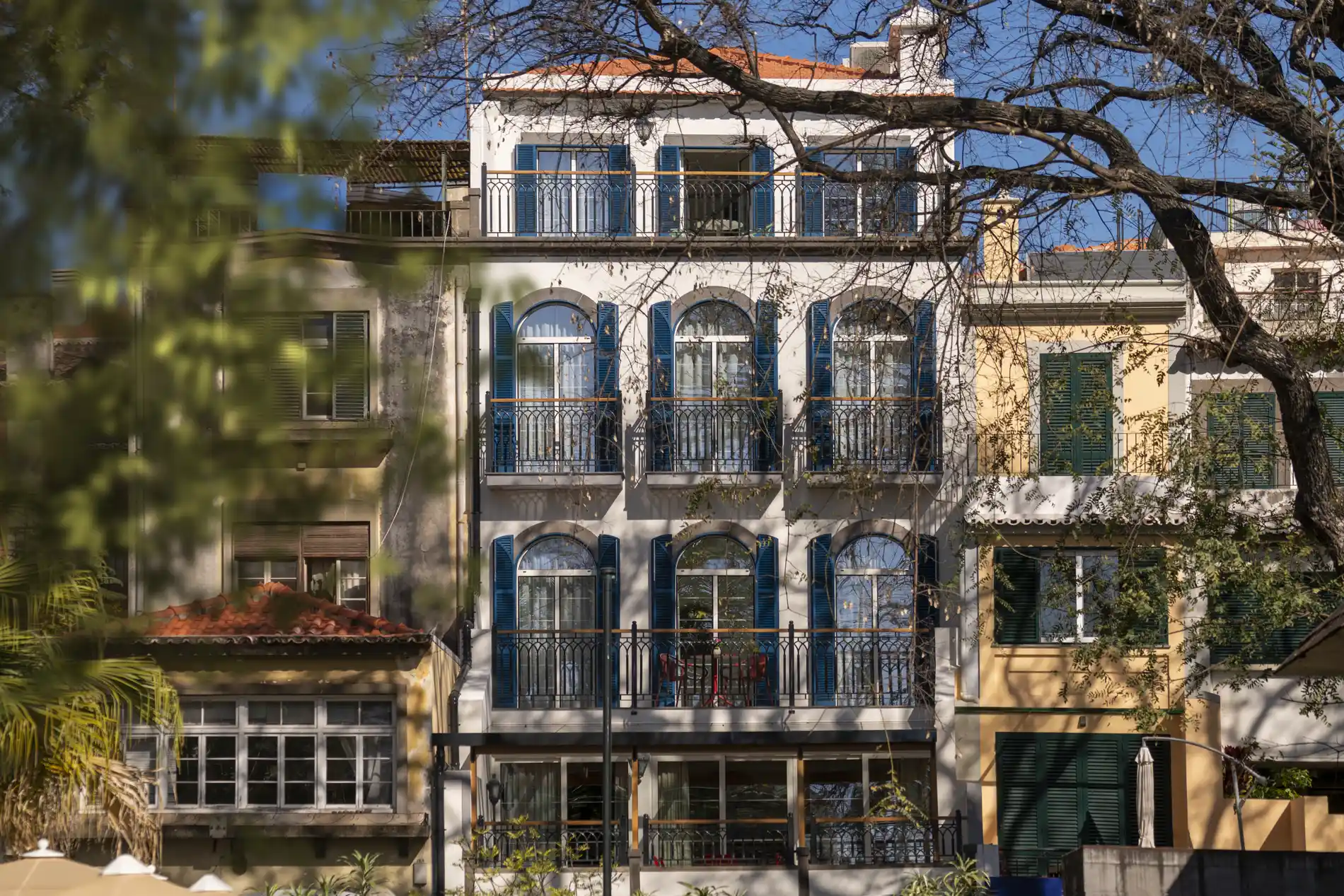 Colorful buildings with blue shutters and balconies, viewed through trees.