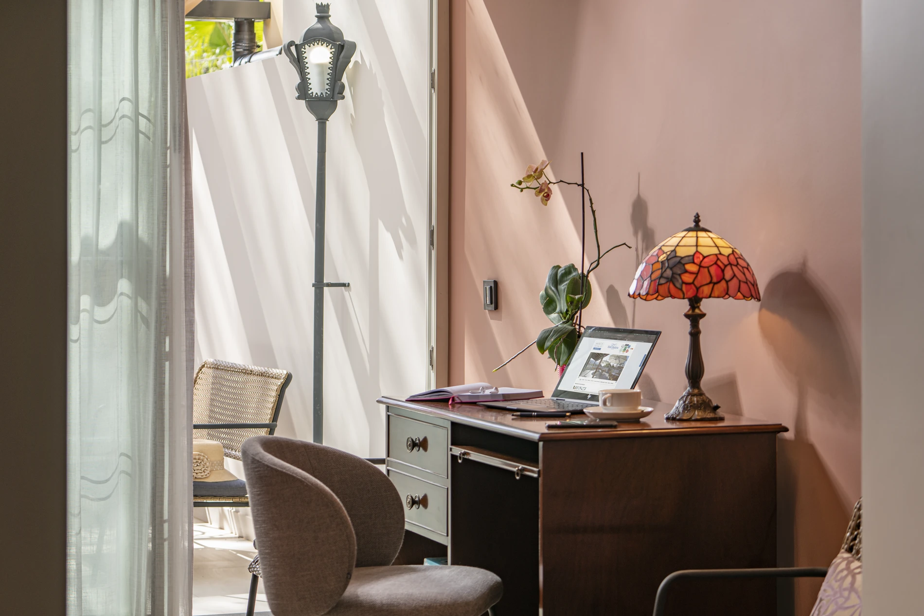 Cozy workspace with a desk, laptop, lamp, and chair by a sunlit window.