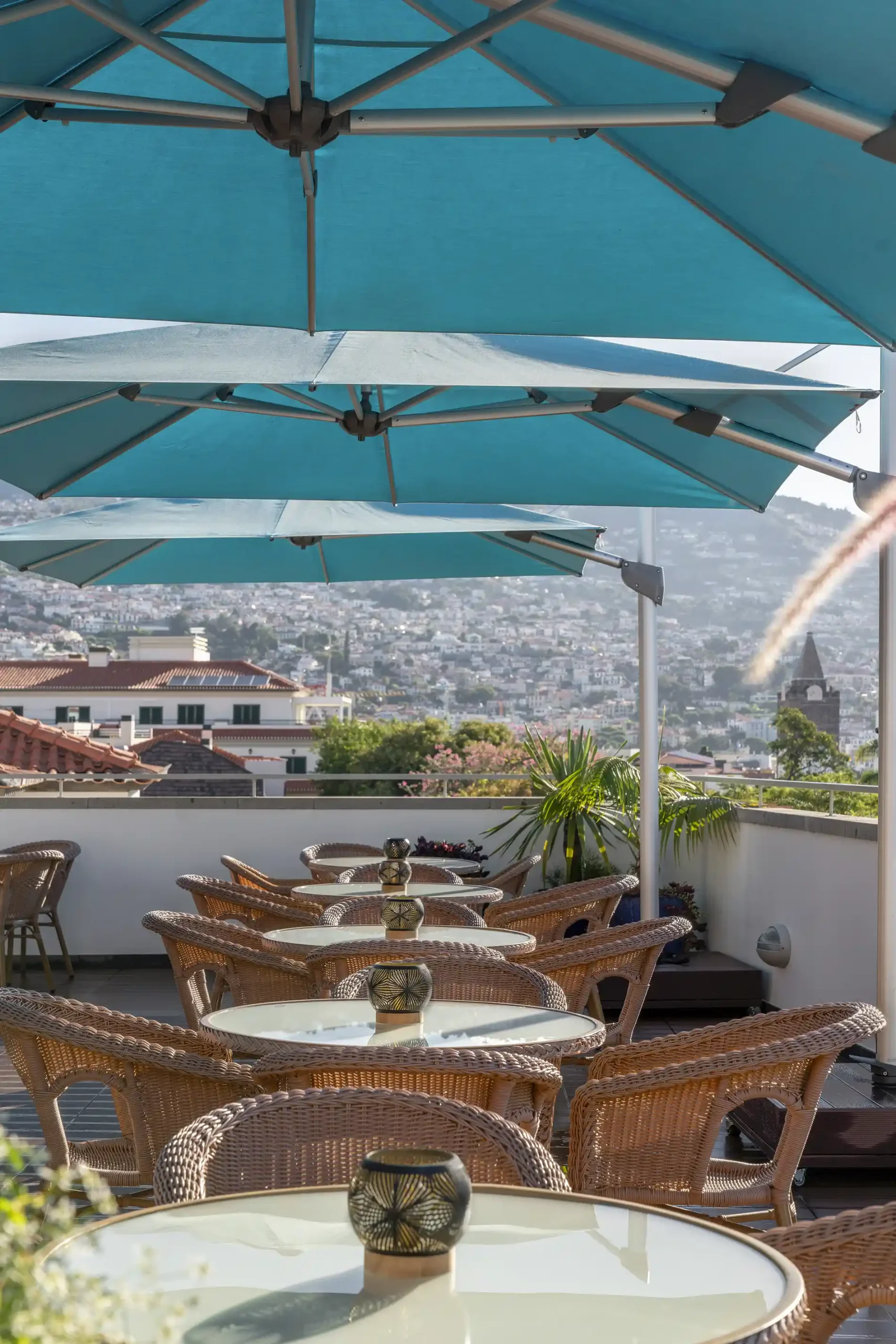 Rooftop patio with wicker chairs, tables, and blue umbrellas overlooking a city.