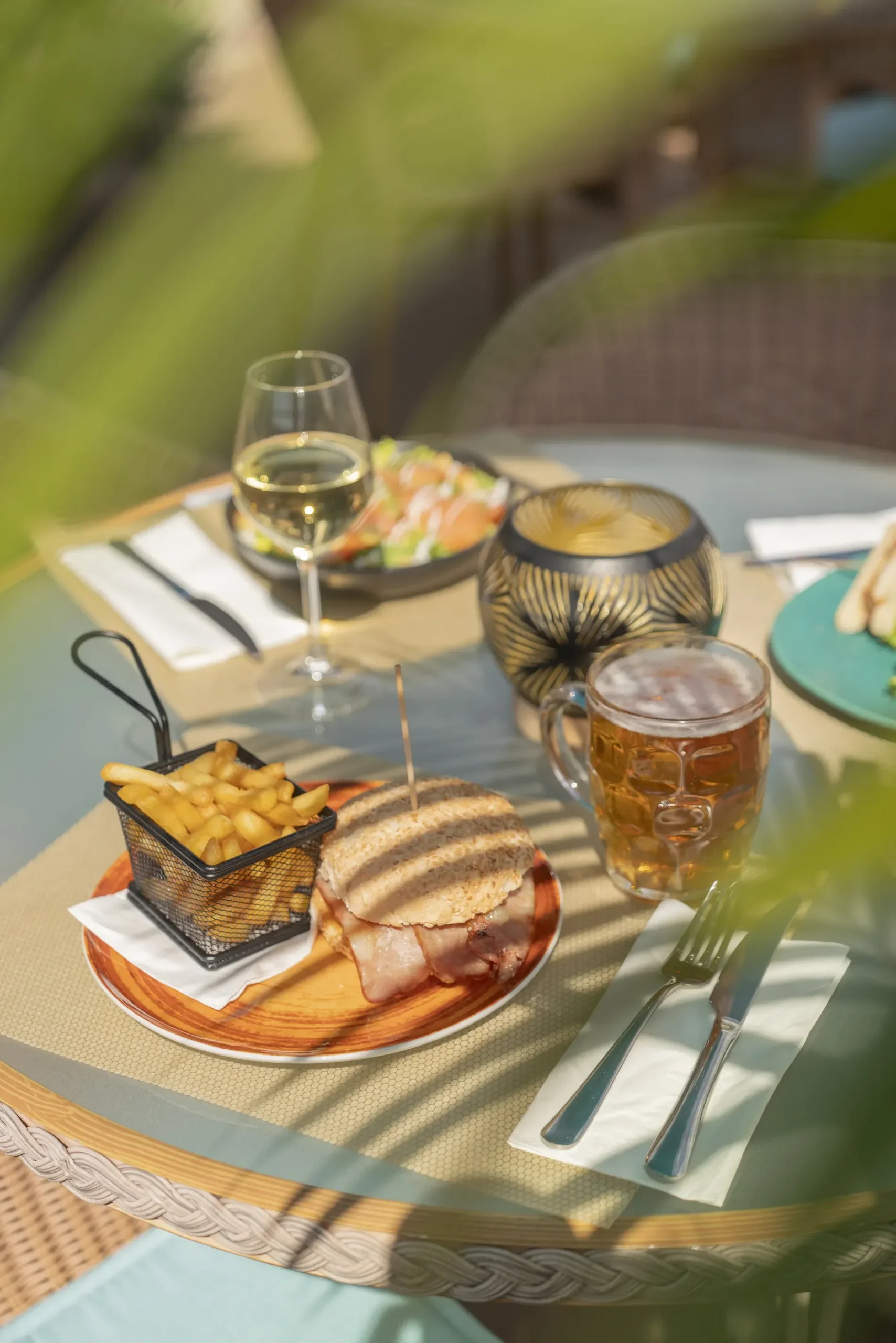 Table with fries, sandwich, beer, wine, and candle under dappled sunlight.