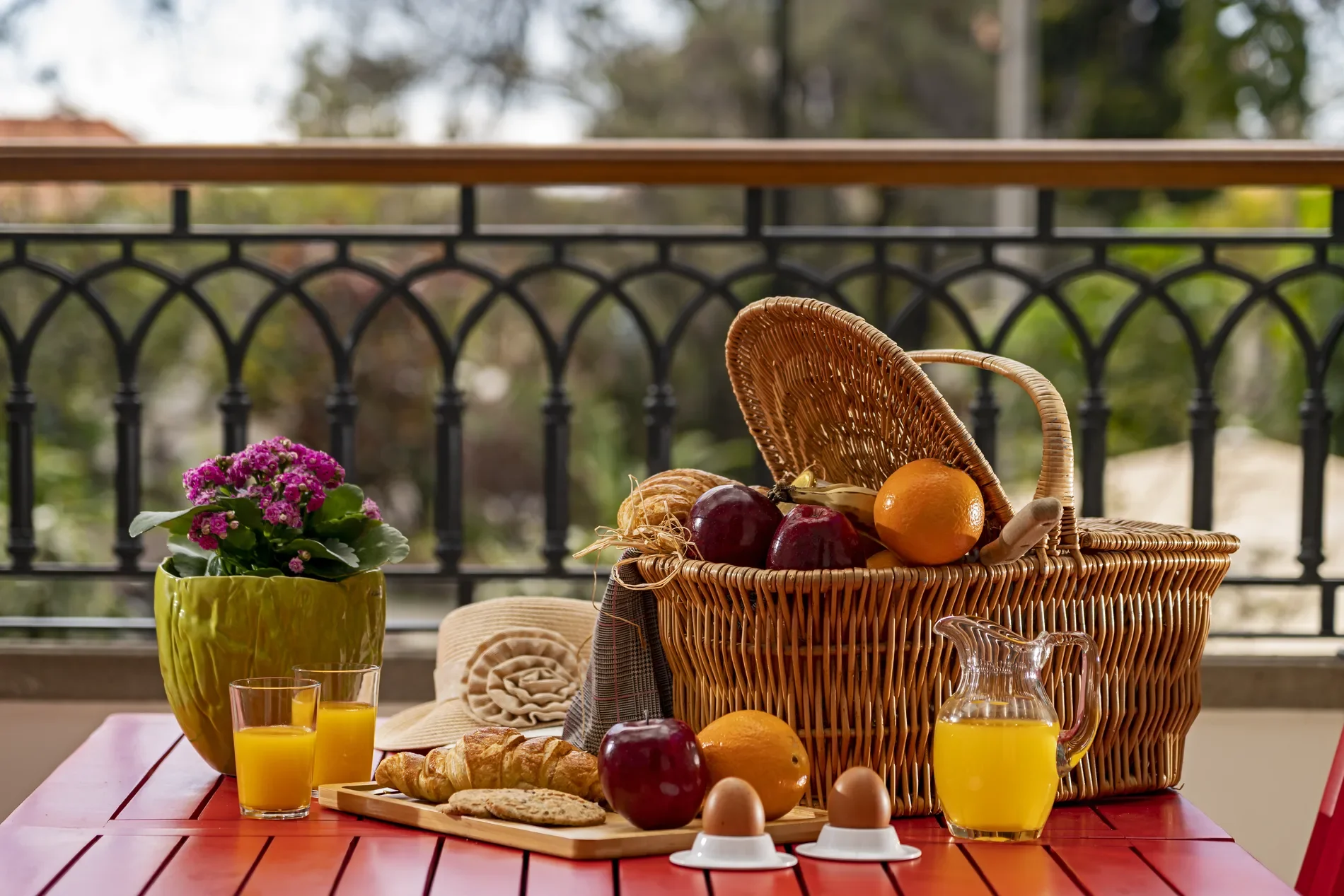 Breakfast items and basket on a red table with flowers and juice.