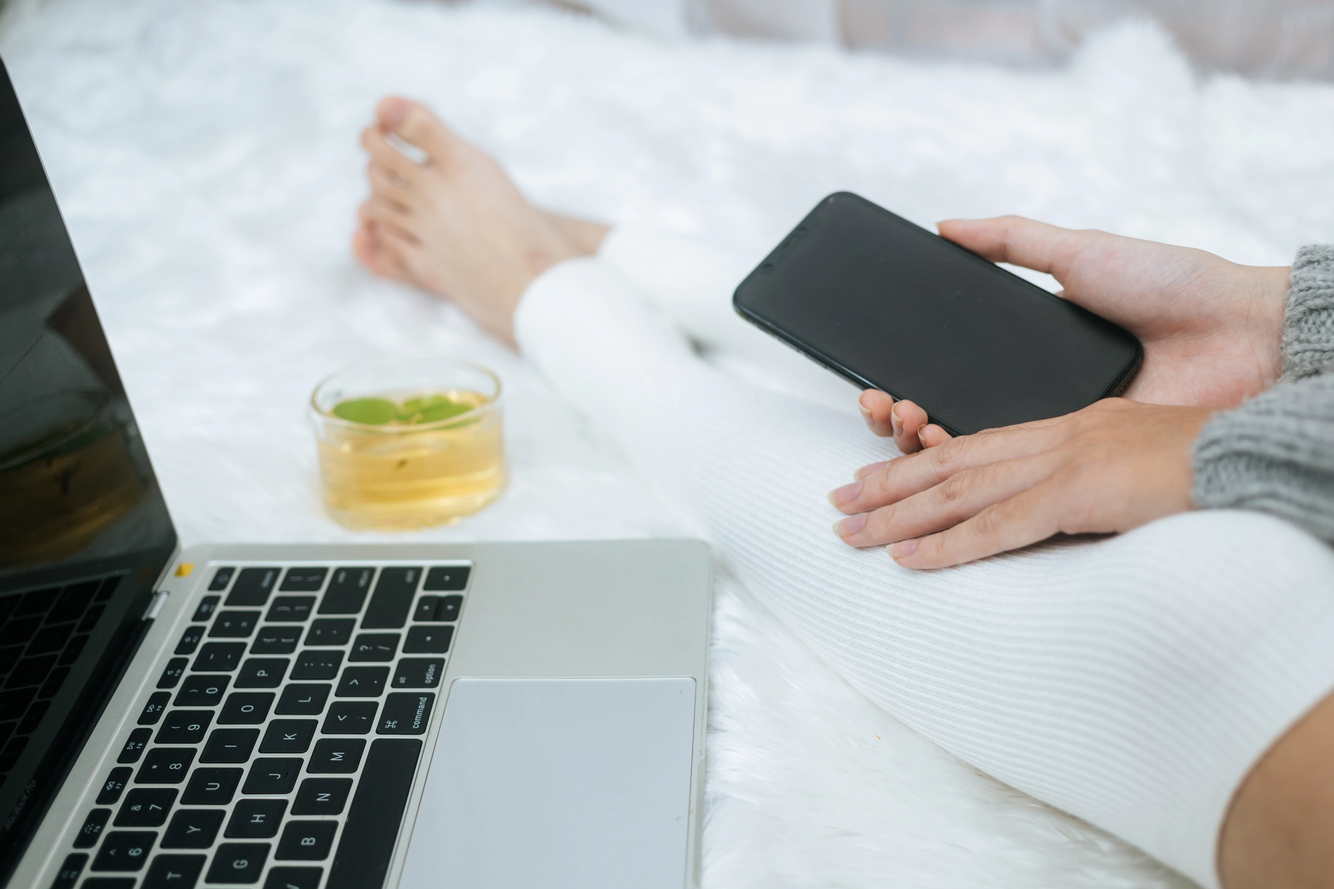 Person using a phone and laptop with tea on a white bed.