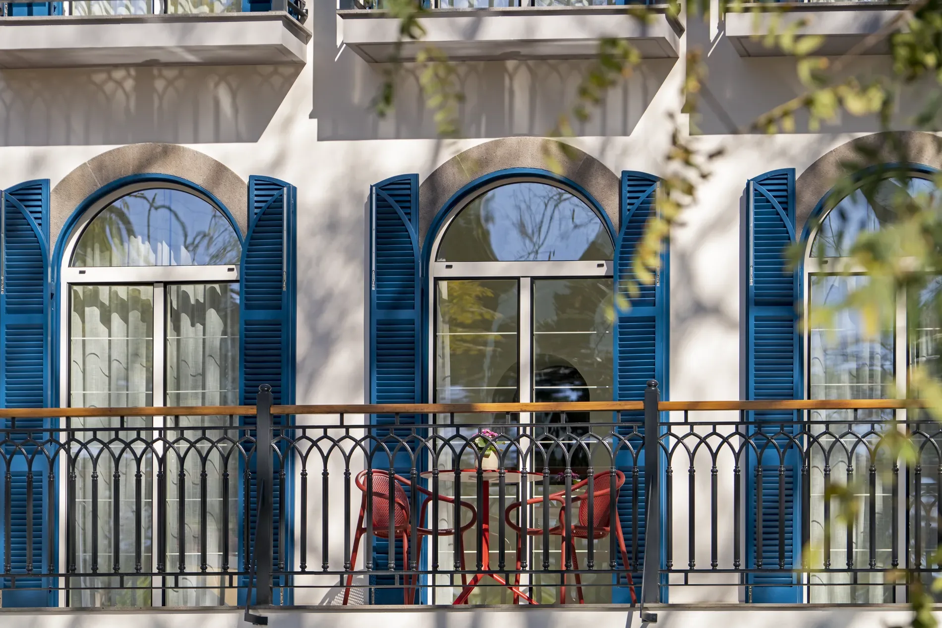 Blue shutters, arched windows, balcony with red chairs and a table.