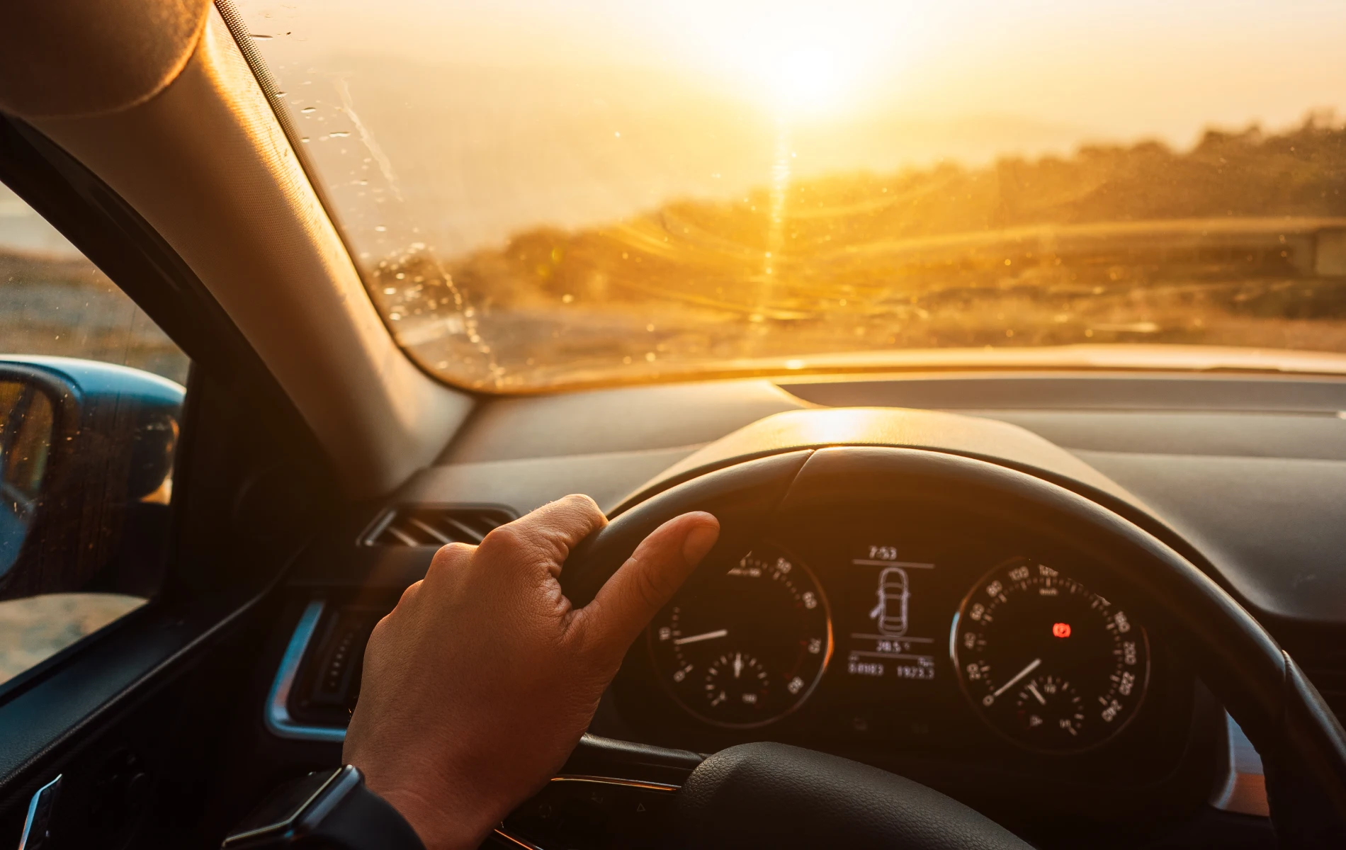 Hand on steering wheel, driving into sunset, view through windshield.