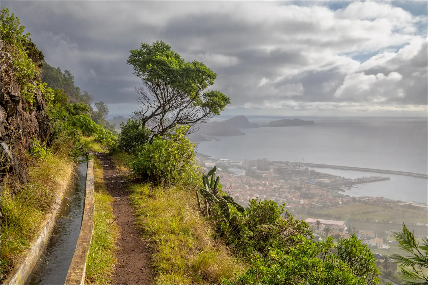 Cliffside path under cloudy sky overlooking ocean and town below.