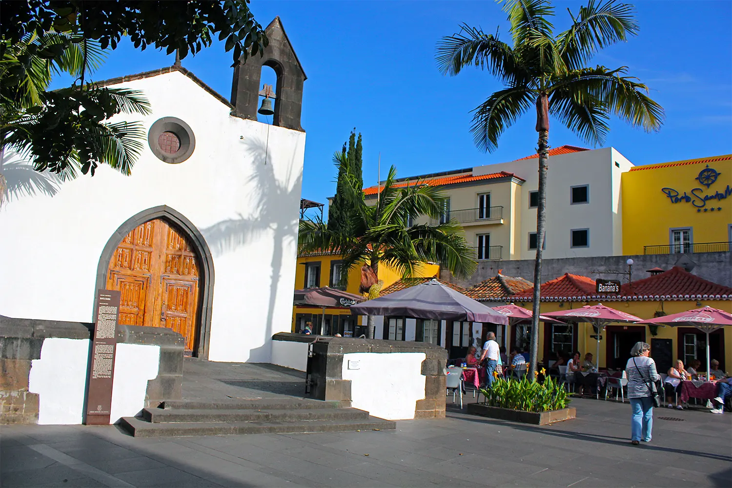 White church and nearby cafe with umbrellas, palm trees, and people outdoors.