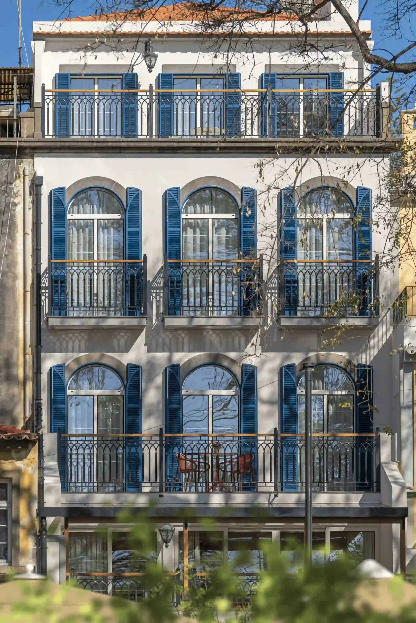 White house with blue shutters and balconies, trees in front, sunny day.