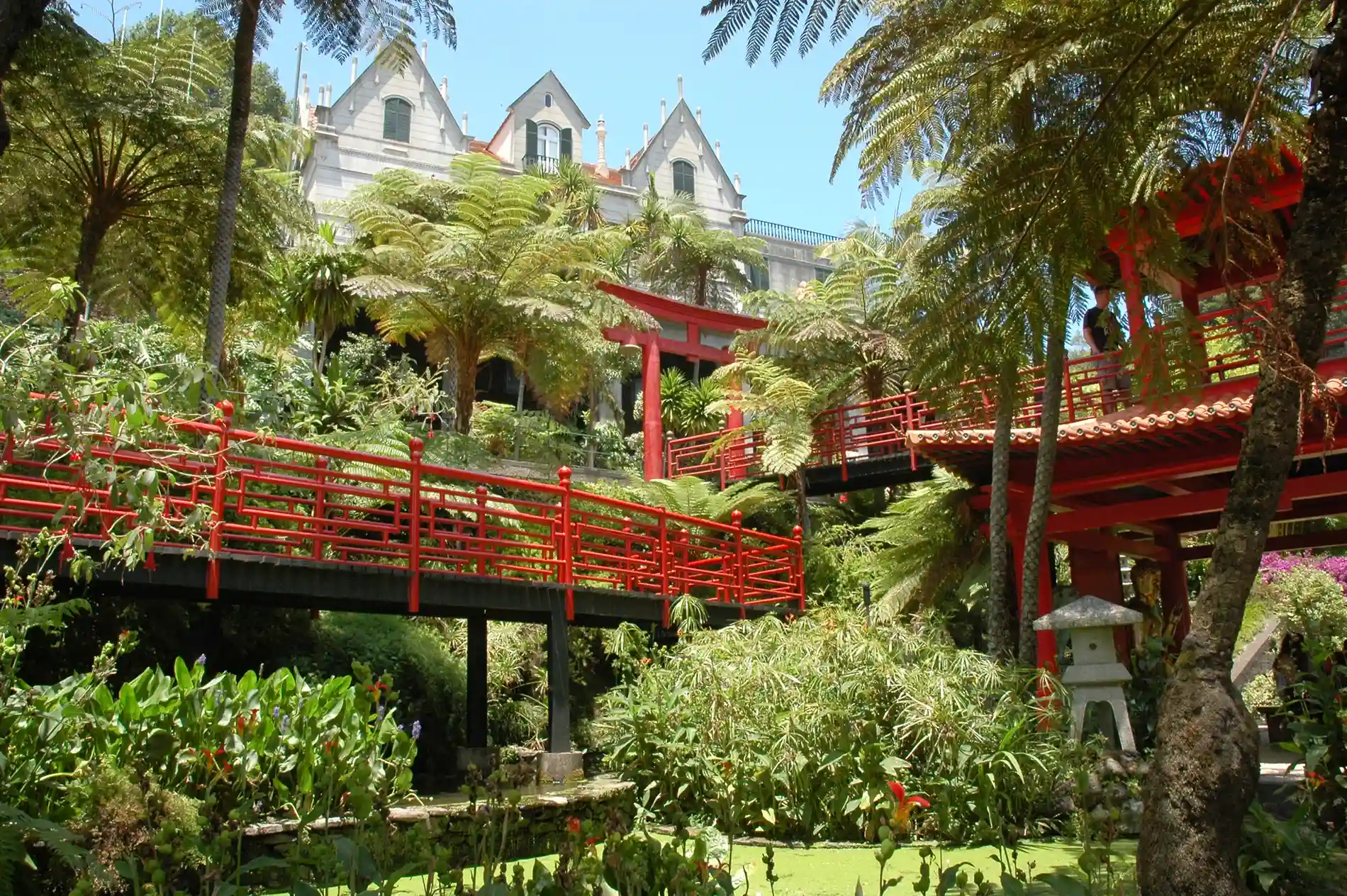 Red bridge in a lush garden with trees and a traditional building in the background.