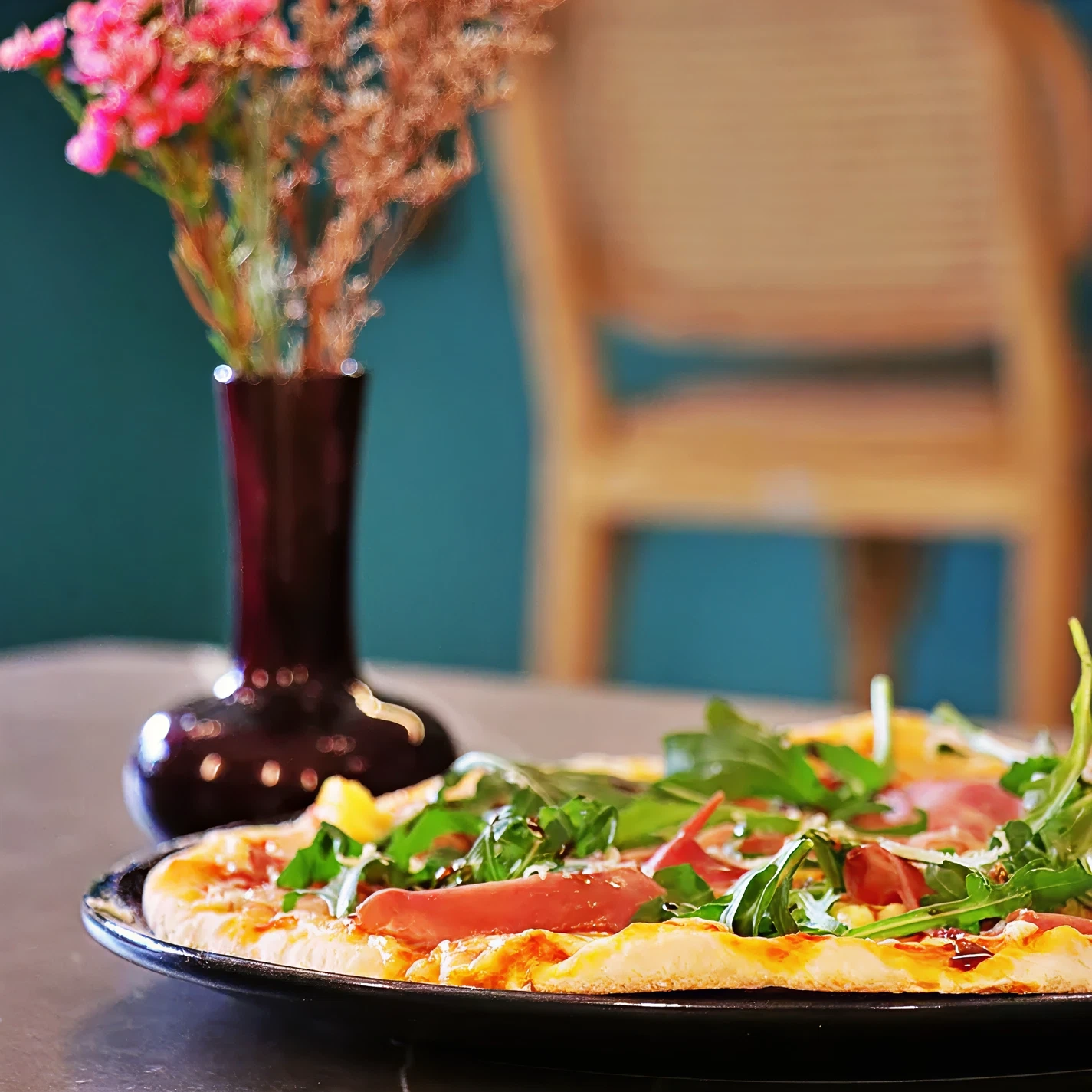 Pizza with arugula and meat next to a vase of flowers on a table.