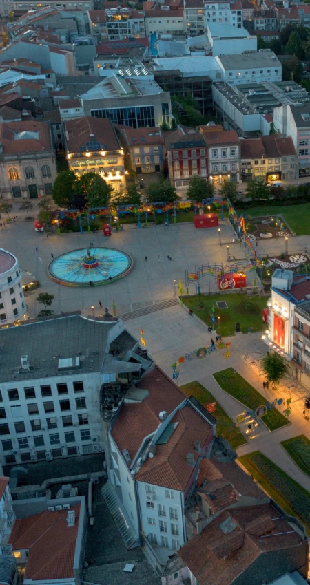 Vista a&eacute;rea de uma pra&ccedil;a com fontes, edif&iacute;cios hist&oacute;ricos e cal&ccedil;ada iluminada.