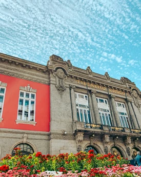 Fachada de teatro antigo sob c&eacute;u azul com nuvens, flores na frente.