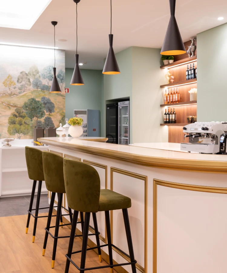 Balc&atilde;o de bar com bancos verdes, lumin&aacute;rias pendentes e prateleira de garrafas.