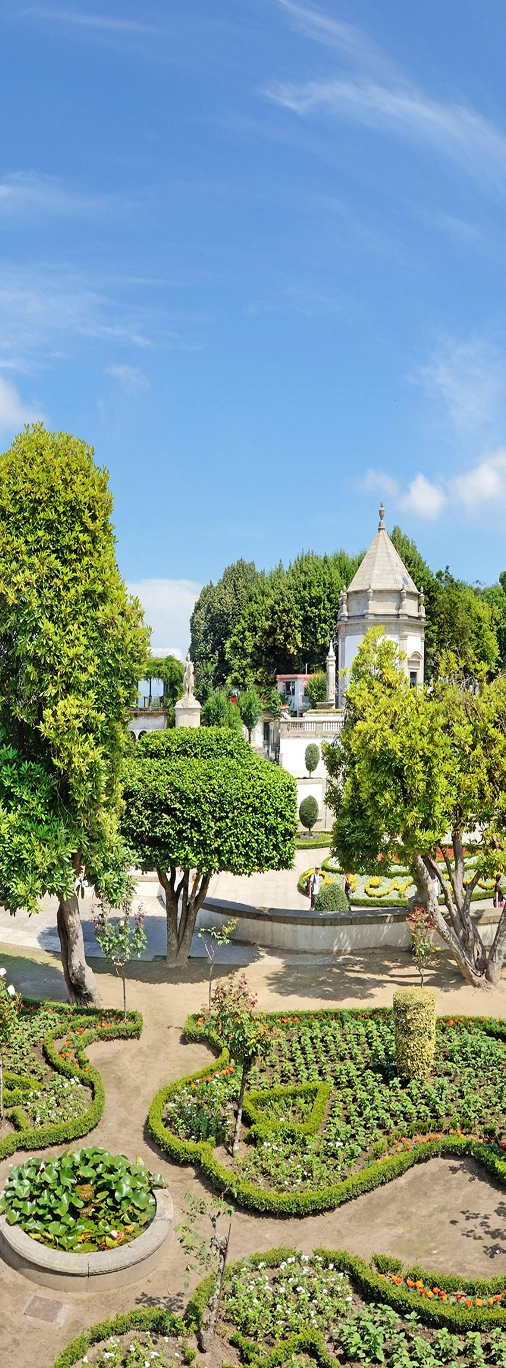 Jardim com canteiros verdes e um edif&iacute;cio hist&oacute;rico ao fundo.