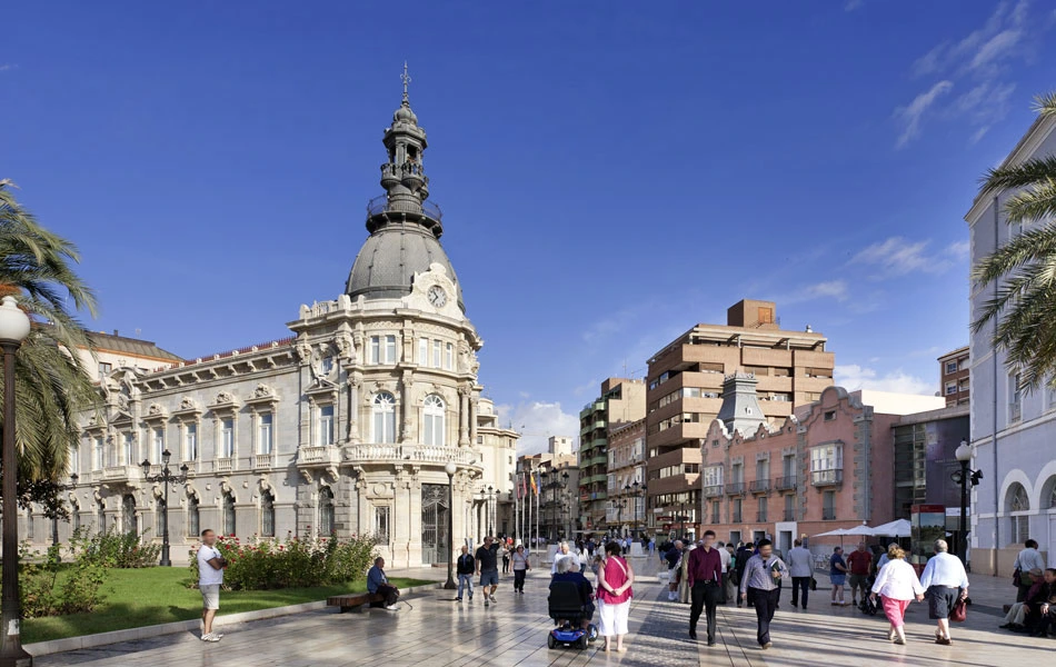 Plaza concurrida con edificio hist&oacute;rico y cielo azul brillante.