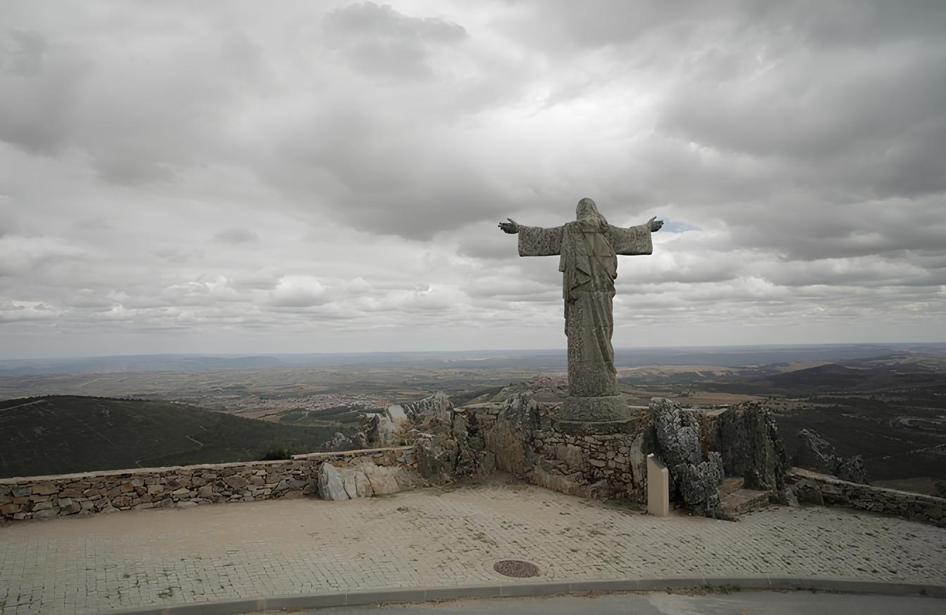 Est&aacute;tua com bra&ccedil;os abertos em meio a uma paisagem montanhosa sob c&eacute;u nublado.