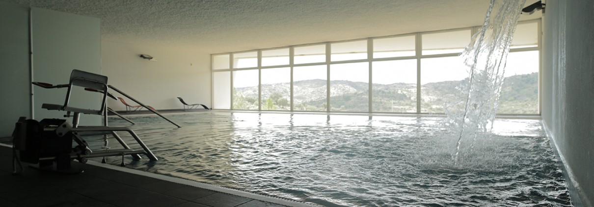 Piscina coberta com vista para colinas atrav&eacute;s de janelas amplas e cascata de &aacute;gua.