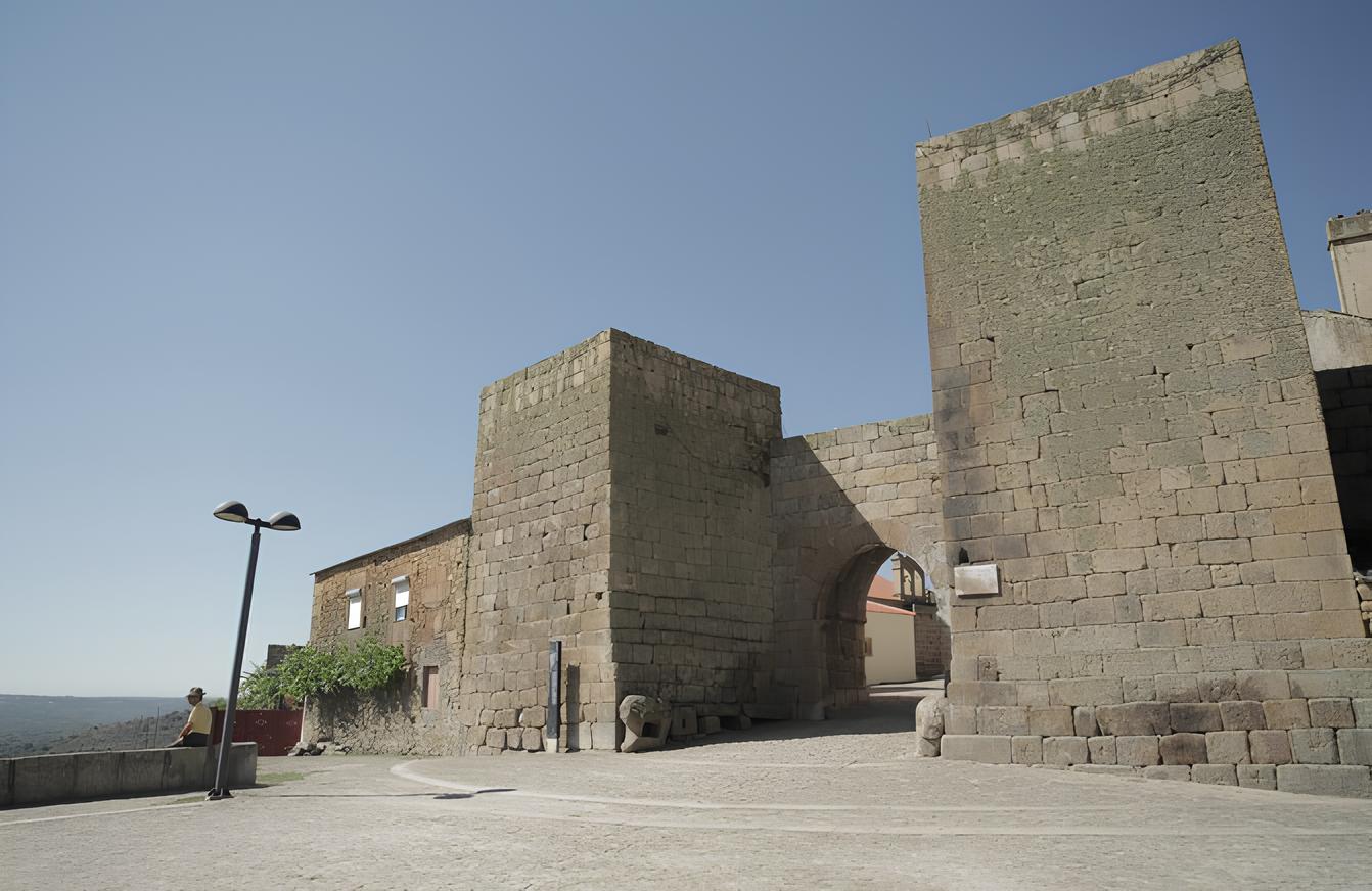 Porta antiga de pedra sob c&eacute;u azul claro, com uma pessoa em p&eacute; pr&oacute;ximo a um corrim&atilde;o.