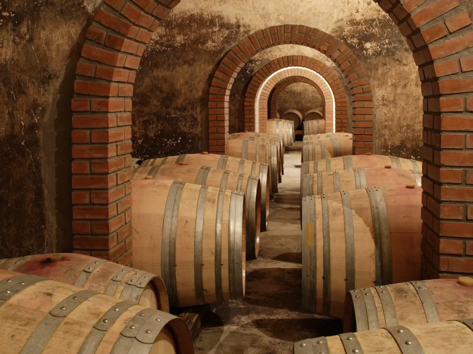 Adega com barris de vinho em um corredor com arcos de tijolos.