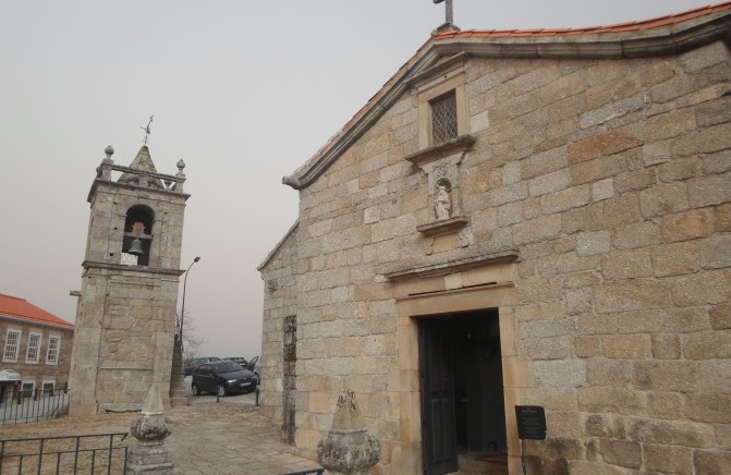 Igreja de pedra com campan&aacute;rio ao lado e carro estacionado ao fundo, c&eacute;u nublado.